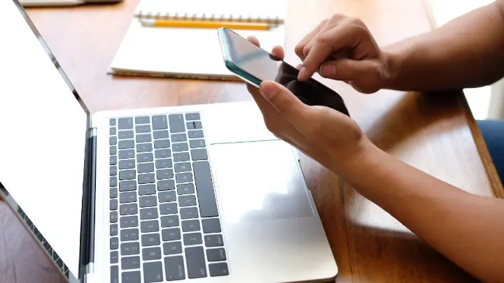 Person scrolling on smart phone with open laptop in front of them